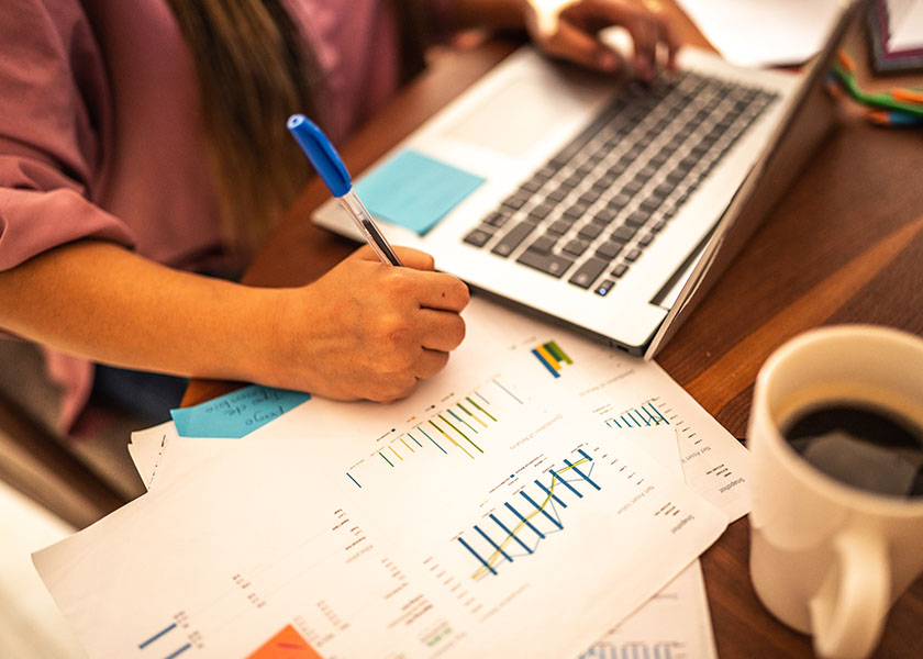 A woman drinking coffee while working at a desk scattered with papers, eyeglasses, and a calculator to determine how to minimize tax savings on his retirement savings.