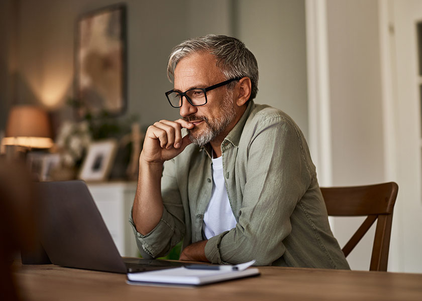 A mature man sitting at the dining room table researching the costs of healthcare in retirement on a laptop.